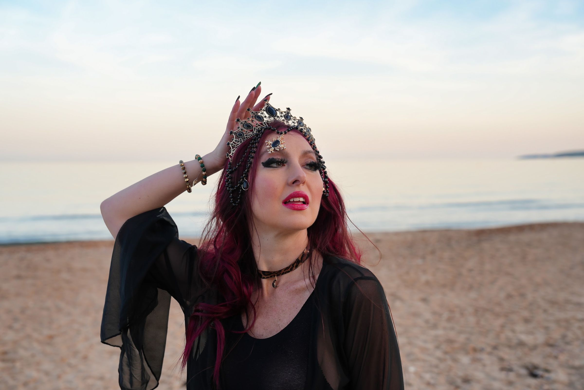 Yulia in a jewelled crown and sheer black sleeves on the shore at dusk