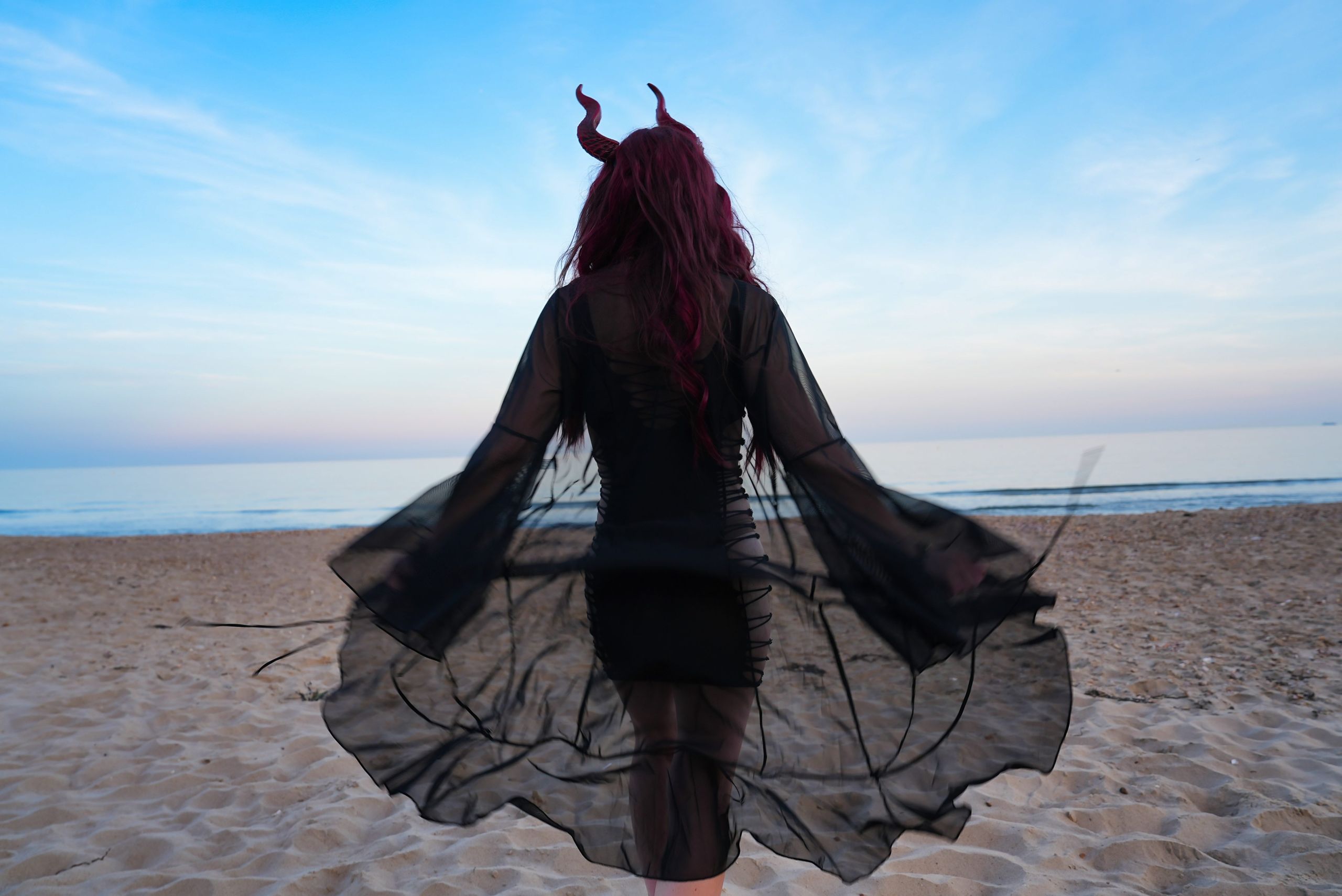 A figure with long red hair and horns walks towards the sea at dusk, black chiffon sleeves billowing