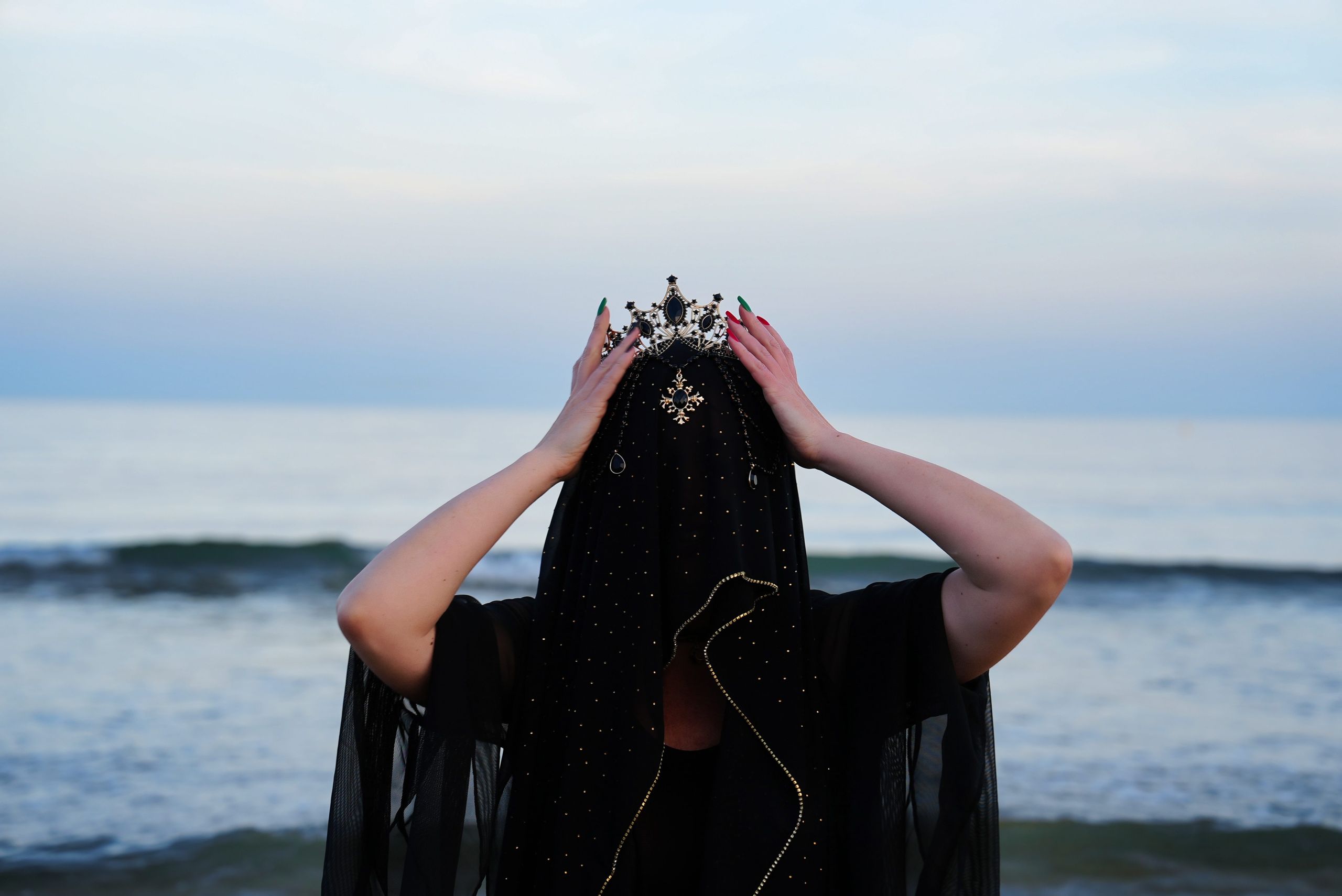 A figure in a black star-flecked veil lifting a jewelled crown at the shoreline at dusk